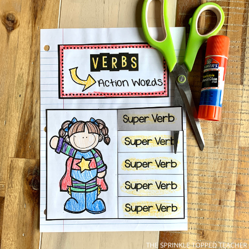 This page of the interactive parts of speech notebook is about verbs. It says that verbs are action words and have an image of a superhero. The students write five super verbs under the flaps of the sheet. Scissors and glue stick lie on the page.