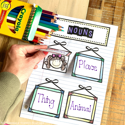 This page shows nouns as person, place, thing and animal. A person hand lifts the person flap to reveal a picture of a little girl with a pink bow. There is a box of colored pencils nearby the interactive parts of speech notebook.