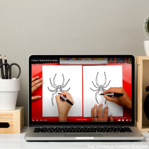 A child practicing drawing a black widow spider on a laptop, sitting on a desk. cup with pens and scissors. 
